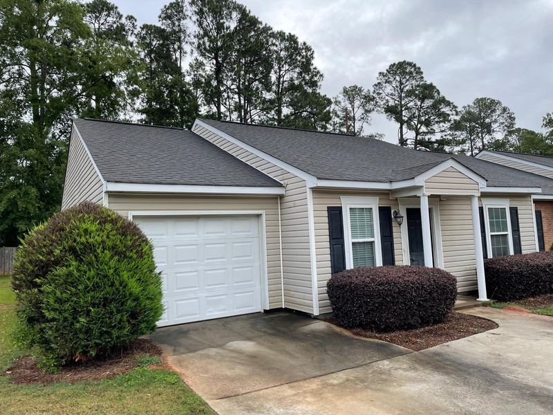 Beige home with black shutters, a covered entry and a one-car garage, under tall pines.