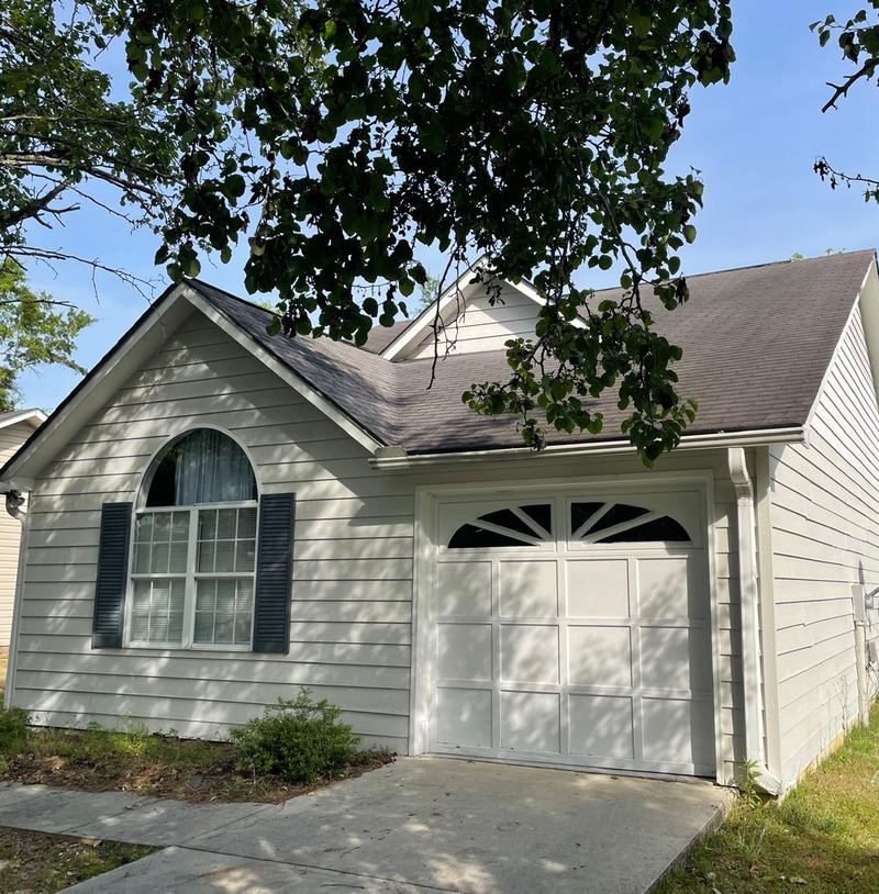 Light gray home with an arched window and a one-car garage, in the shade of a large tree.